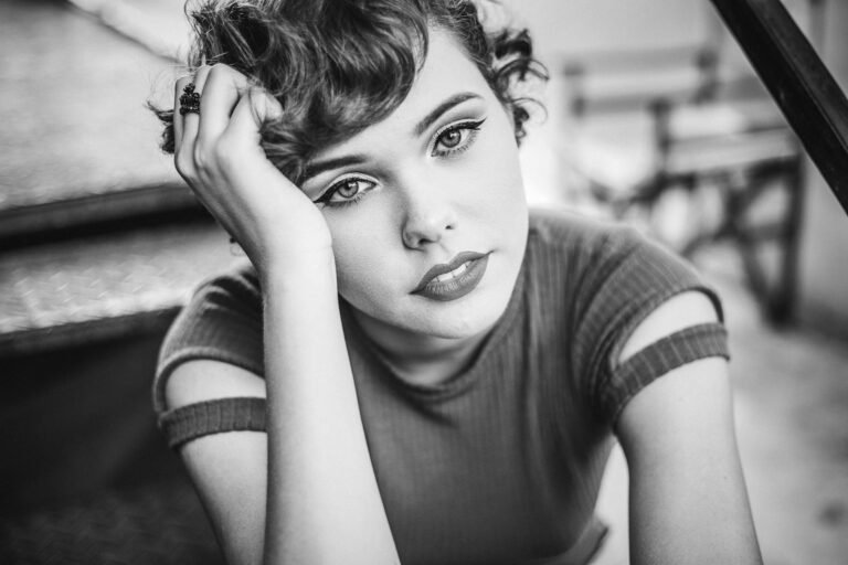 Retrato em preto e branco de uma mulher jovem com cabelo curto ondulado, apoiando o rosto na mão e olhando fixamente para a câmera.